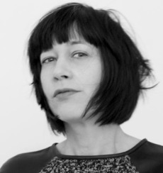 Schwarzweißfoto einer Person mit kurzen, dunklen, zerzausten Haaren, die ein gemustertes Oberteil trägt und vor einem schlichten Hintergrund leicht zur Seite blickt.