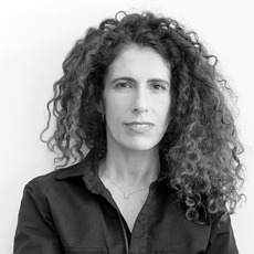 Eine Frau mit lockigem Haar, einem dunklen Hemd mit Knöpfen und einer Halskette steht vor einem schlichten hellen Hintergrund und blickt direkt in die Kamera.