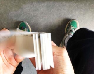 Eine Person blättert durch einen kleinen Stapel weißer Karten, während sie auf einem grauen Boden steht und grün-weiße Turnschuhe trägt.