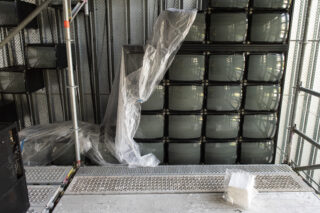 In einer Industriehalle mit Metallwänden und Gerüst ist ein Stapel alter Fernseher teilweise mit durchsichtiger Plastikfolie abgedeckt.