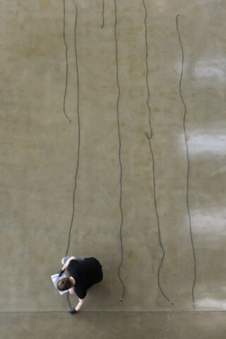 Eine Person in Schwarz ordnet oder inspiziert mehrere lange schwarze Kabel, die in parallelen Linien auf einem großen, glatten, hellbraunen Boden verlegt sind.
