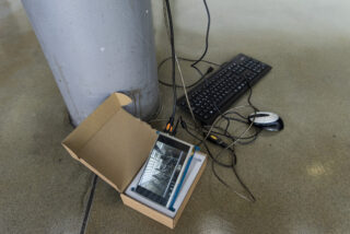 Tastatur, Maus und Kabel liegen neben einer Säule auf dem Boden. In einem offenen Karton befindet sich ein Tablet mit einer schwarz-weißen Architekturdarstellung.