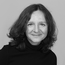 Eine Frau mit schulterlangem, welligem Haar und einem schwarzen Rollkragenpullover lächelt leicht vor einem schlichten, hellen Hintergrund.