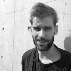 Ein Mann mit Bart und kurzen Haaren steht vor einer Betonwand und blickt in die Kamera. Das Bild ist schwarz-weiß.