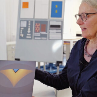 Eine Person mit Brille hält ein geometrisches Kunstwerk in einem gut beleuchteten Raum hoch, während im Hintergrund eine Tafel mit Designmustern zu sehen ist.