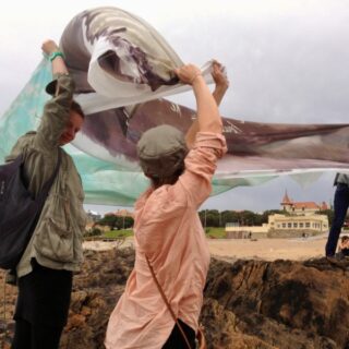 Zwei Personen auf felsigem Boden halten ein großes Stofftuch, möglicherweise ein Banner oder eine Flagge, hoch, während es im Wind flattert. Im Hintergrund sind ein Strand und Gebäude zu sehen.