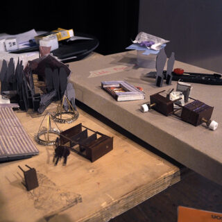 Auf einer Tischplatte sind Miniaturmöbel, ausgeschnittene Figuren und Architekturmodelle sowie Bastelwerkzeuge und Materialien ausgestellt, die rund um den Arbeitsbereich angeordnet sind.