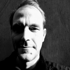 Schwarzweißfoto eines Mannes, der in die Kamera blickt, wobei die Hälfte seines Gesichts im Schatten liegt und die andere Hälfte beleuchtet ist, vor einem schlichten strukturierten Hintergrund.