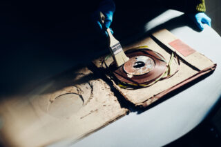 Eine Person mit blauen Handschuhen reinigt mit einer Bürste ein beschädigtes Tonbandgerät in einer alten, abgenutzten Hülle auf einem Tisch.