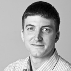 Ein Mann mit kurzen dunklen Haaren und kariertem Hemd blickt in die Kamera und lächelt leicht vor einem schlichten Hintergrund.