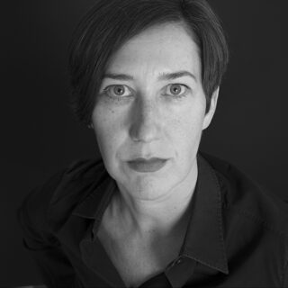 Eine Person mit kurzen dunklen Haaren und ernstem Gesichtsausdruck blickt in die Kamera. Sie trägt ein dunkles Button-up-Hemd vor einem dunklen Hintergrund.