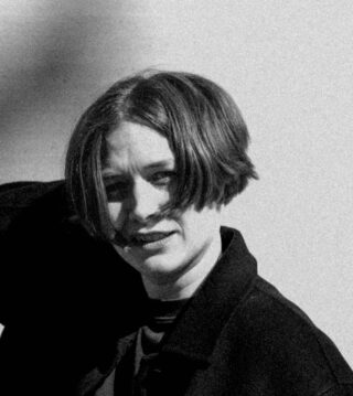 Black and white photo of a person with short hair and a dark jacket, looking at the camera with one hand raised behind their head against a plain background.