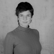 Eine Person mit kurzen Haaren, Brille und geripptem Rollkragenshirt posiert vor einem schlichten, hellen Hintergrund. Das Bild ist schwarz-weiß.