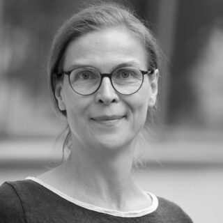 A woman with glasses and tied-back hair looks at the camera and smiles slightly. The photo is in black and white.