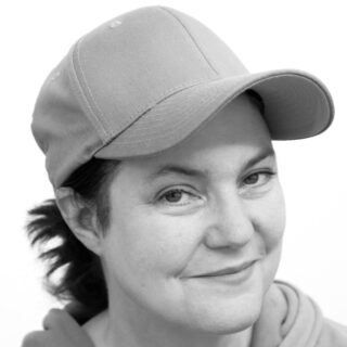 Eine Frau mit Baseballkappe und Kapuzenpullover blickt mit einem leichten Lächeln in die Kamera. Das Bild ist schwarz-weiß.