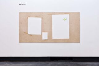 Eine Holztafel mit drei weißen, papierähnlichen Formen, von denen eine an den Ecken mit Klebeband befestigt ist, an einer Wand angebracht; in der oberen linken Ecke steht der Text „Hello Illusion!“.