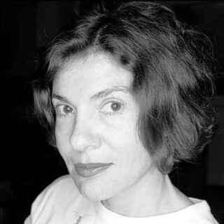 Schwarzweißfoto einer Frau mit kurzem, welligem Haar, die ein helles Oberteil trägt und mit neutralem Gesichtsausdruck direkt in die Kamera blickt.