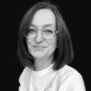 Eine Frau mittleren Alters mit schulterlangen dunklen Haaren, die eine runde Brille und ein helles Oberteil trägt, lächelt sanft vor einem schlichten schwarzen Hintergrund.