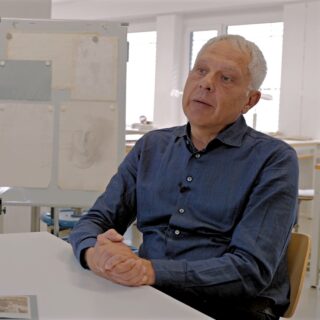 Ein älterer Mann mit grauem Haar sitzt an einem Tisch, die Hände gefaltet, in einem blauen Hemd. Hinter ihm steht eine Tafel, an die Papiere und Skizzen geheftet sind.