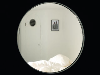 Ein rundes Fenster mit Blick auf einen weißen Raum mit geschlossener Tür, einem gerahmten Bild an der Wand und einem ungemachten Bett mit weißen Laken.