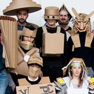 Eine Gruppe von Menschen in verschiedenen Kostümen aus Pappe, darunter Hüte, Masken, Rüstungen und Accessoires, steht zusammen und blickt in die Kamera.