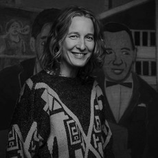 Eine lächelnde Frau mit welligem Haar steht vor einem Wandgemälde mit zwei Männern in Anzügen.