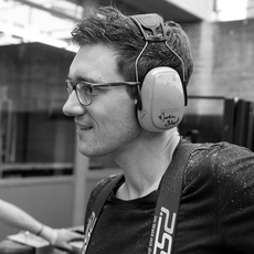 Ein Mann mit Brille und Ohrenschützern steht in einem Innenbereich mit Blick nach links. Er trägt ein T-Shirt und Hosenträger und befindet sich in einer werkstattähnlichen Umgebung.