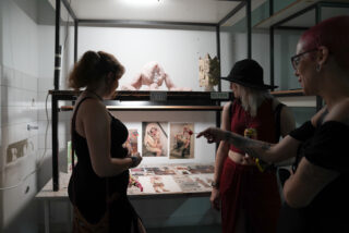 Drei Personen betrachten Kunstwerke und Fotografien, die auf einem Tisch und einem Regal in einem Studio oder einer Galerie ausgestellt sind.