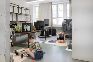 Zwei Personen arbeiten auf dem Boden eines hellen Raums an Kunstprojekten, umgeben von Materialien, Regalen, Papieren und Schautafeln.