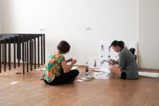 Zwei Personen sitzen auf einem Holzboden, umgeben von Papieren und Künstlerbedarf, und arbeiten in einem hellen, minimalistischen Raum an Zeichnungen oder Skizzen.