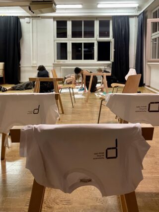 In einem Raum hängen mehrere weiße T-Shirts über Holzständern, während im Hintergrund zwei Personen auf dem Boden in der Nähe von offenen Fenstern und Kunstbedarf arbeiten.