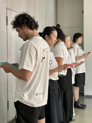 Fünf Personen in weißen T-Shirts stehen in einer Reihe im Innenbereich, lesen in bunten Broschüren und blicken nach links.