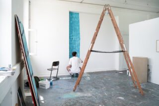 Eine Person sitzt auf dem Boden und malt ein blaues Gitter auf eine weiße Wand in einem geräumigen, gut beleuchteten Kunstatelier mit einer Holzleiter und verstreuten Materialien.