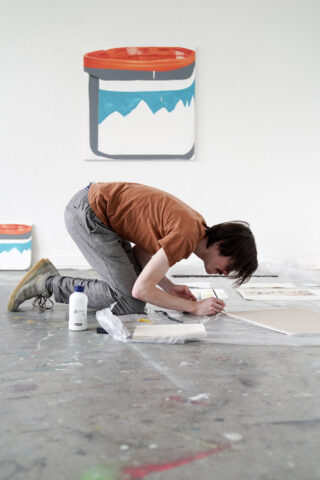 Eine Person kniet auf dem Boden und arbeitet an einer Leinwand. In der Nähe stehen Künstlerutensilien. Im Hintergrund hängt an der Wand das Bild einer Farbdose.