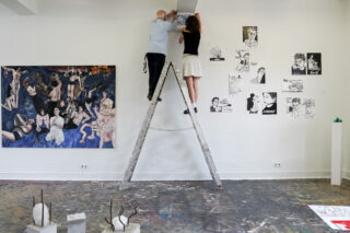 Zwei Personen stehen auf gegenüberliegenden Seiten einer Leiter und befestigen etwas an der Wand in einem Kunstatelier, umgeben von verschiedenen Zeichnungen, Gemälden und Skulpturen.