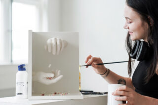 Eine Frau bemalt eine weiße Skulptur aus zwei Händen, von denen eine Pillen in die andere wirft. Auf dem Tisch neben ihr liegen Malutensilien und eine Flasche mit der Aufschrift „ALCOHETE ETHANOL“.