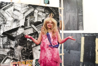 Eine Person in einem rosa Kleid, einer blauen Schärpe und einer Krone steht mit ausgestreckten Armen vor einem schwarz-weißen Architekturgemälde, umgeben von anderen Kunstwerken.