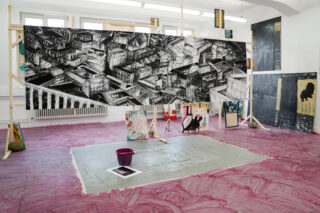 Ein Kunstatelier mit einer großen schwarz-weißen Stadtlandschaftszeichnung auf einem Holzrahmen, umgeben von bunten Gemälden und einem lila Eimer auf einem rosa gestrichenen Boden.