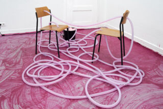 Auf einem rosafarbenen Boden stehen zwei Holzstühle, die durch lange, geschwungene rosafarbene Röhren verbunden sind, die sich zwischen ihnen und um sie herum winden und verwickeln.