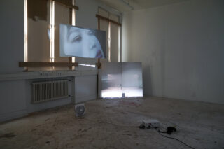 Ein Raum mit zwei Videoprojektionen: eine zeigt die Nahaufnahme eines Gesichts, die andere eine entfernte Gestalt; auf dem Boden liegen freiliegende Kabel und ein Ventilator.