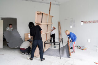 Fünf Personen montieren oder bewegen große Holz- und Metallkonstruktionen in einem weißen Raum mit hängenden Objekten und verstreuten Materialien.