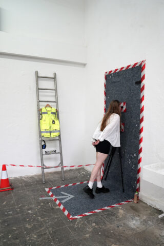 Eine Frau blickt durch eine grau gestreifte Barriere, die an einer Wand befestigt ist. In der Nähe stehen in einem weißen, kargen Raum eine Leiter, eine Warnweste und ein Verkehrsleitkegel.