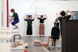 Vier Personen arrangieren und hängen gerahmte Kunstwerke an einer weißen Wand in einem Kunststudio auf, das mit Kisten, Zubehör und Möbeln gefüllt ist.