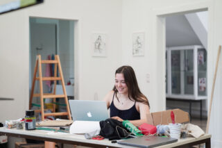 Eine junge Frau sitzt an einem unordentlichen Tisch und arbeitet an einem Laptop in einem hellen Raum. Im Hintergrund stehen Künstlerbedarf und eine Staffelei.