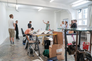 Eine Gruppe von Menschen versammelt sich in einem überfüllten Atelier mit Schreibtischen, Kunstbedarf und Pappkartons. Eine Person gestikuliert, während die anderen zuschauen.