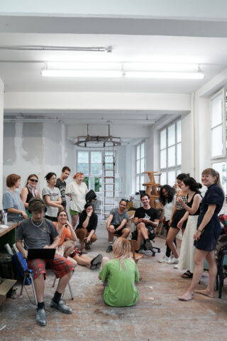 Eine Gruppe von Menschen trifft sich und unterhält sich in einem hellen, geräumigen Kunstatelier mit großen Fenstern, verstreuten Stühlen und sichtbaren Kunstutensilien.