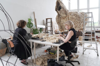 Zwei Personen arbeiten an einem Tisch und verarbeiten Wolle. Überall liegen Wollstücke herum, im Hintergrund ist eine große, mit Wolle bedeckte Struktur zu sehen. Das Studio ist hell.