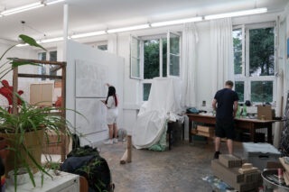 Zwei Personen arbeiten in einem gut beleuchteten Kunstatelier. Eine malt Text auf eine große Leinwand, die andere sitzt an einem unordentlichen Schreibtisch. Verschiedene Malutensilien und Pflanzen füllen den Raum.