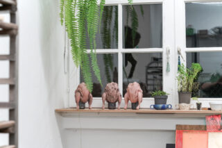 Drei rohe ganze Hühner liegen kopfüber auf einem Fensterbrett, umgeben von Topfpflanzen und Küchenutensilien, darüber hängen Farne vor einem weißen Fenster.