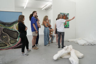 Eine Gruppe von Menschen steht in einer Kunstgalerie und hört einer Person zu, die auf ein buntes Gemälde an der Wand deutet; auf dem Boden stehen Skulpturen.